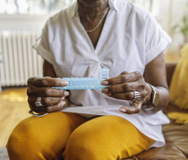 Senior black woman sits on the couch at home and takes medications from a daily pill organizer. Cropped shot does not show the woman